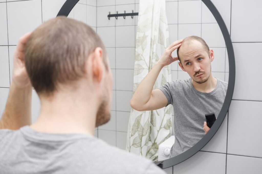 homem de frente para um espelho redondo, aparentando observar sua cabeça com uma expressão preocupada