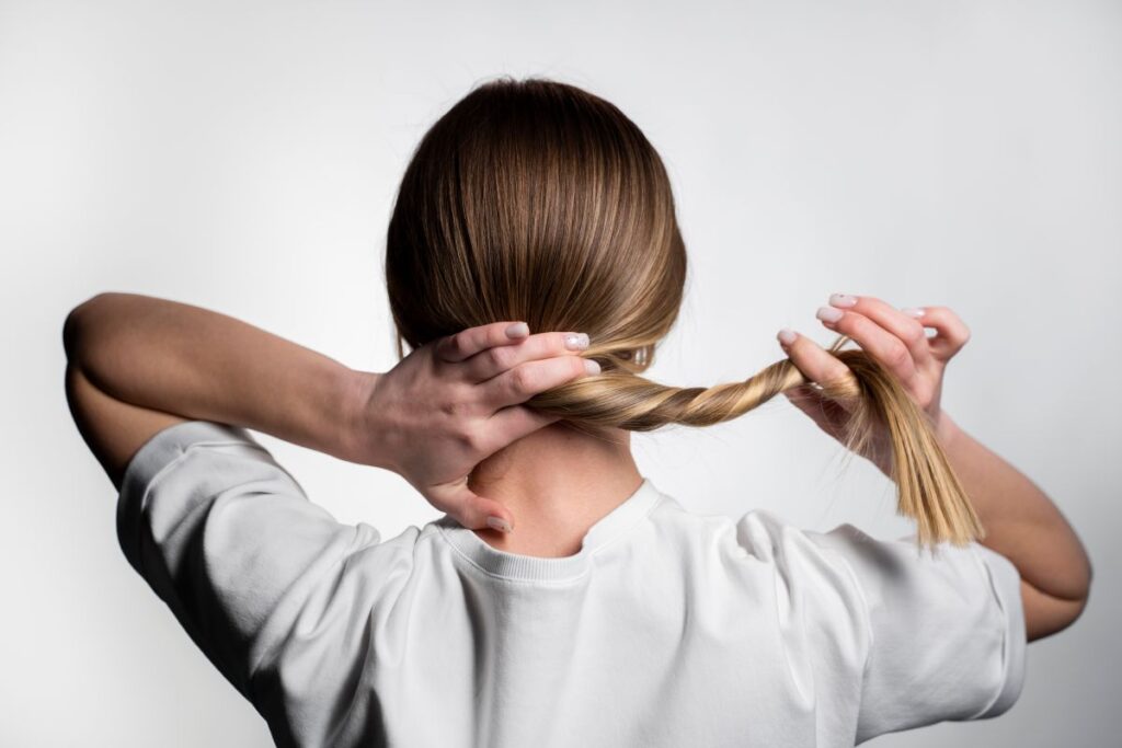 Tratamento para fortalecer o cabelo: x técnicas modernas mulher de costas, vestindo uma camiseta branca, segurando o próprio cabelo loiro e torcendo-o levemente com as mãos.