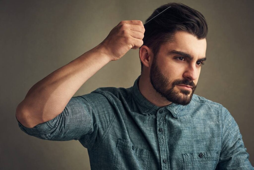 Natural hairline: quais são as características importantes? Homem jovem, de barba cheia e aparência confiante, está penteando os cabelos escuros para trás com um pente fino. Ele veste uma camisa jeans abotoada até o colarinho e olha com foco para o próprio reflexo fora de cena. A imagem destaca sua natural hairline, bem definida e simétrica, evidenciando uma linha capilar saudável e preservada, sem sinais aparentes de entradas ou rarefação.
